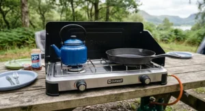 A close-up view of a stainless steel double burner camping stove on a wooden picnic table, featuring a blue enamel kettle on the left hob and a cast iron frying pan on the right.