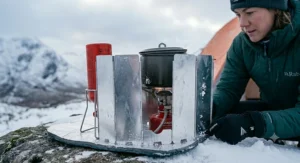 A multi-panel aluminium windshield wrapped tightly around a backpacking stove to protect the flame from wind on a cold winter hillside.