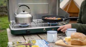 Close-up of a compact folding double burner camping stove, showcasing the burner controls and stainless steel finish.