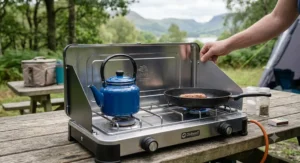 A person adjusting the side windshield of a stainless steel double burner camping stove, which has a blue kettle boiling and a sausage frying in a pan.