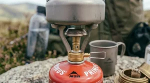 A low-angle close-up of a small metal camping kettle balanced on the serrated pot support arms of a screw top camping stove.